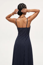 A woman modeling a navy blue midi dress with thin vertical pinstripes, shown from a three-quarters back view against a plain white studio background.