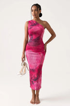 A woman modeling a one-shoulder midi dress with a pink, magenta, and dark purple abstract watercolor pattern, holding a pair of white high-heeled sandals against a plain white studio background.