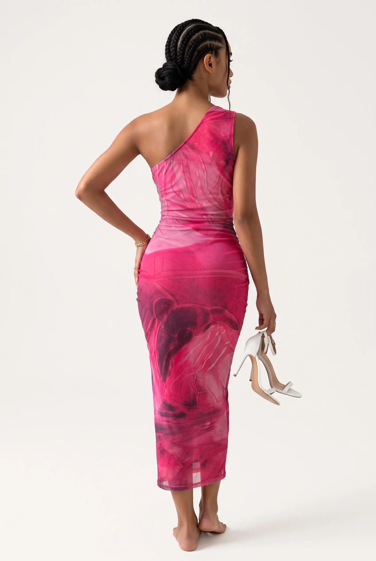 A woman modeling a one-shoulder pink dress with an abstract print, shown from the back against a light gray studio background. She is holding a pair of white high-heeled sandals.