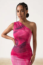 A woman modeling a one-shoulder sheer mesh dress with a vibrant pink and dark purple abstract watercolor pattern, set against a plain off-white studio background.