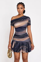A woman modeling a one-shoulder ruched mini dress with a blue, beige, and brown abstract swirl pattern, holding a gold clutch against a plain white background.