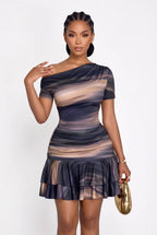 A woman modeling a short-sleeve off-the-shoulder mini dress with a dark blue and beige abstract brushstroke pattern, holding a gold clutch against a plain white background.