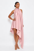 A model wearing a light pink high-low halter dress with a draped, flowing satin-like fabric, posed against a clean white studio background.