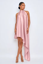 A model wearing a high-neck, sleeveless pink satin dress with an asymmetrical draped hem, paired with clear high-heeled sandals, set against a plain light gray studio background.