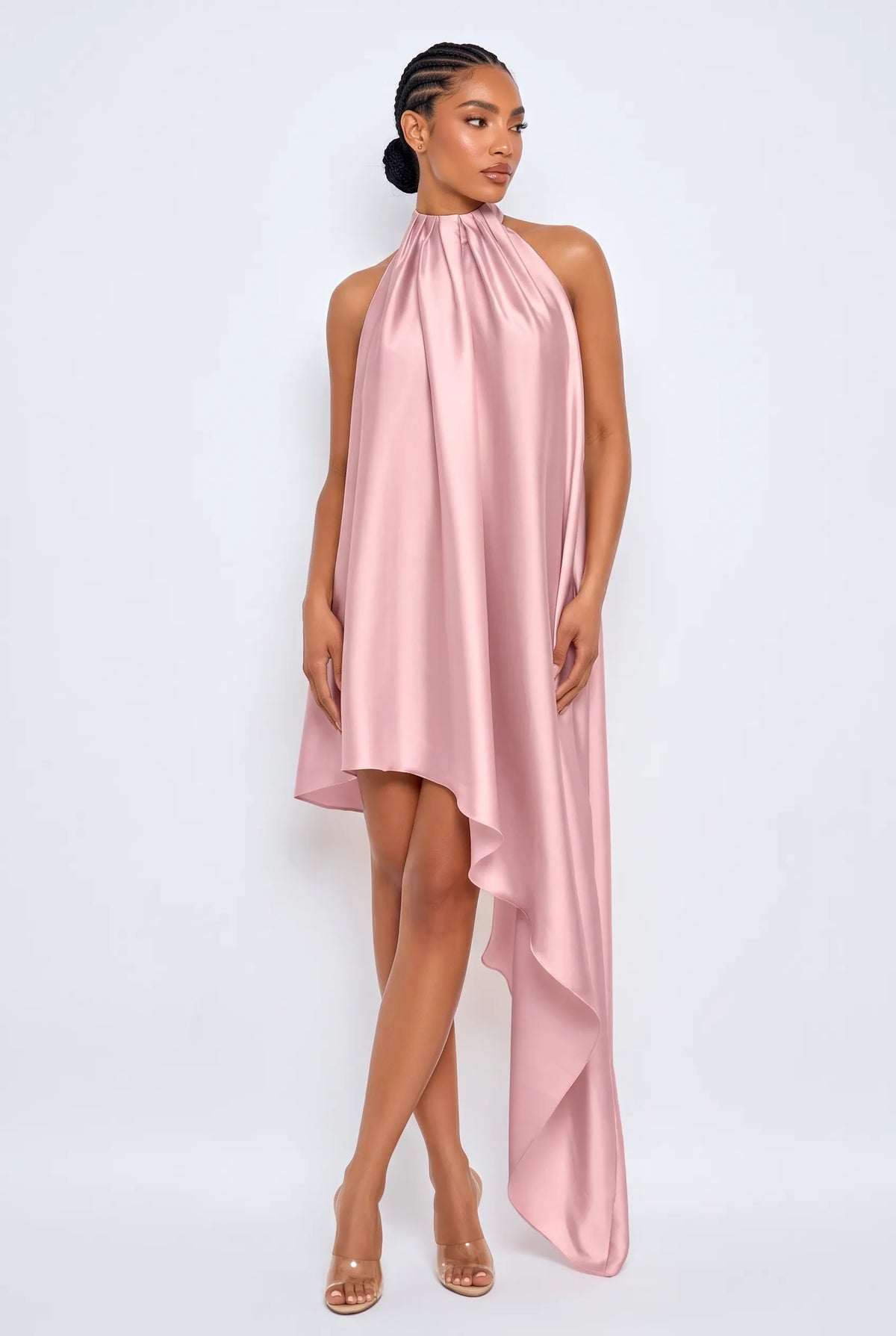 A model wearing a high-neck, sleeveless pink satin dress with an asymmetrical draped hem, paired with clear high-heeled sandals, set against a plain light gray studio background.