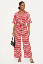 A woman modeling a red and white striped short-sleeve jumpsuit with a tie waist, paired with clear high-heeled sandals and a small black handbag, against a plain white studio background.