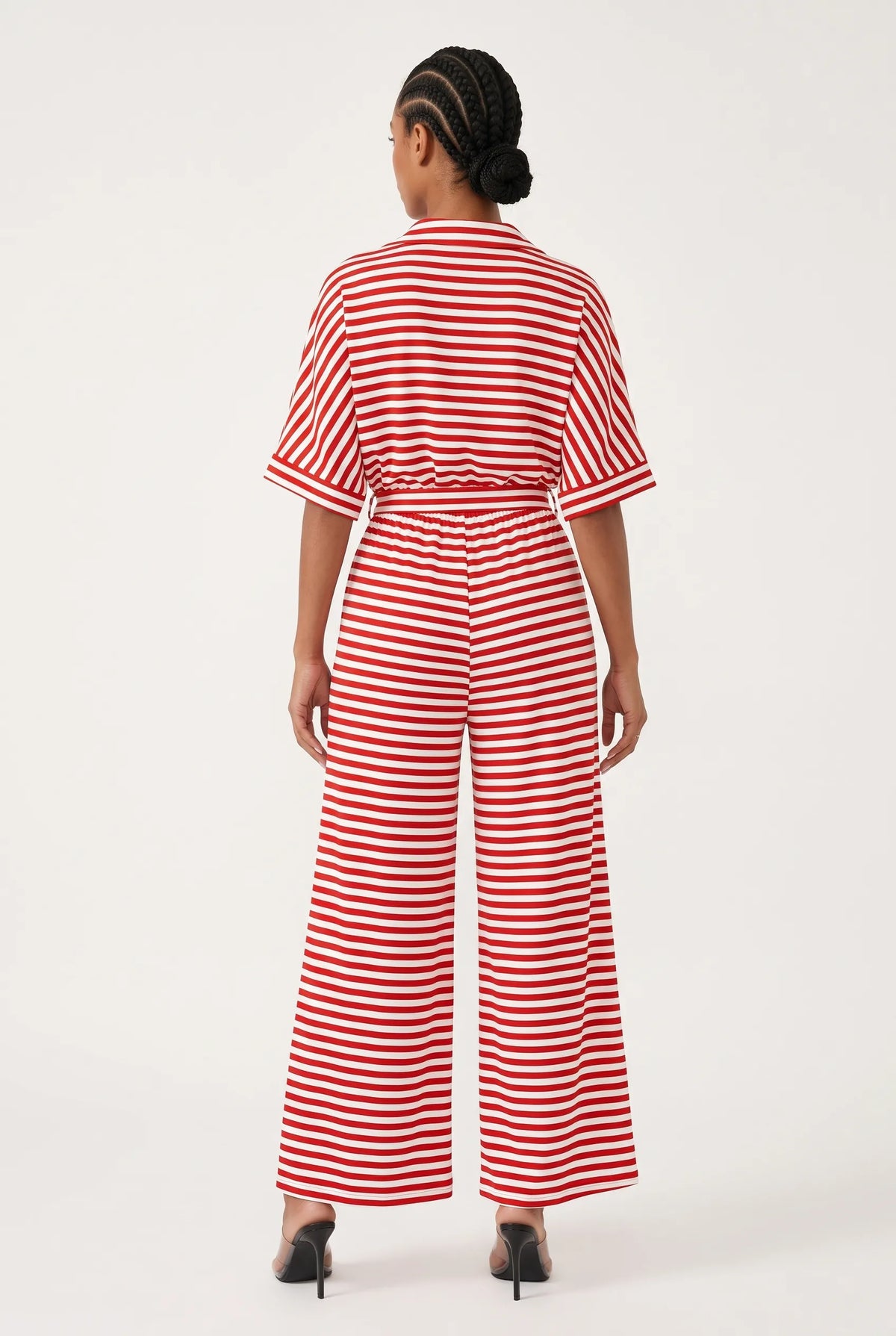 A woman viewed from the back wearing a red and white horizontally striped short-sleeve jumpsuit with a tie waist and wide legs, paired with black heels, against a plain white studio background.