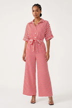 A woman modeling a red and white horizontally striped short-sleeve jumpsuit with a button-up front, collar, and matching tie belt at the waist, photographed against a plain light background.