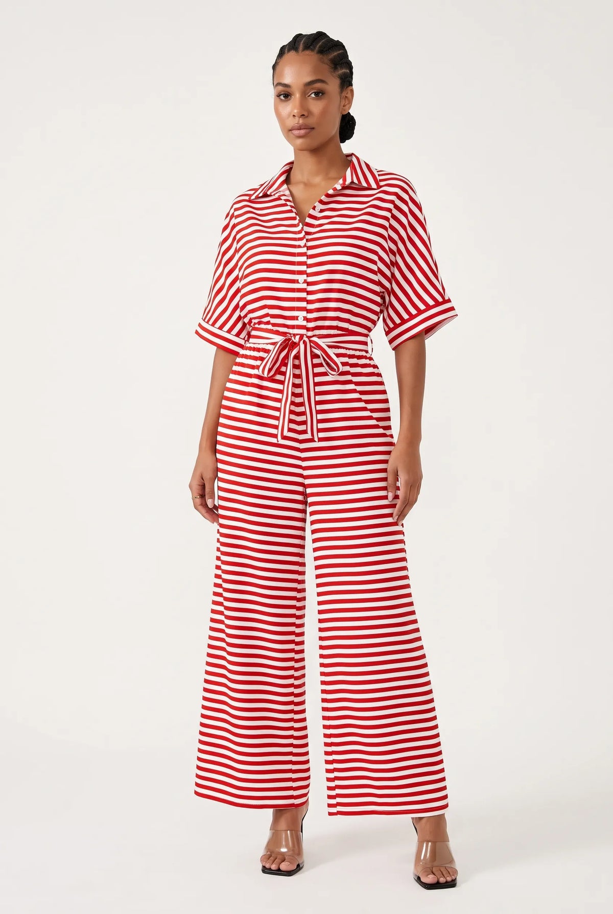 A woman modeling a red and white horizontally striped short-sleeve jumpsuit with a button-up front, collar, and matching tie belt at the waist, photographed against a plain light background.