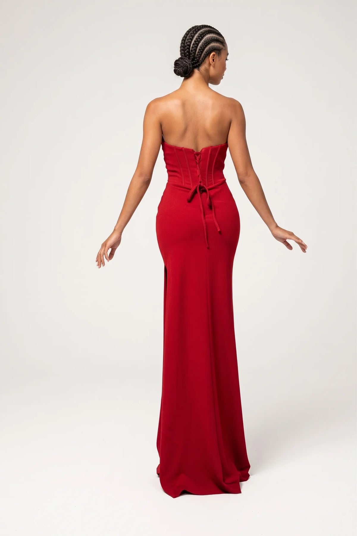 A woman modeling a strapless red floor-length gown with a ruched bodice, draped skirt, and front slit, photographed against a plain light gray studio background.