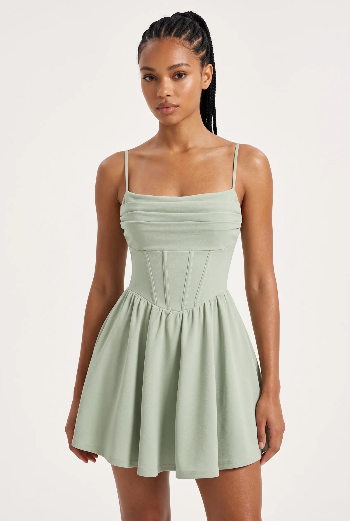 A woman modeling a sage green spaghetti-strap mini dress with a ruched bodice and flared skirt against a plain light background.