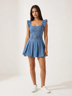 Model wearing a short-sleeved denim mini dress is crafted from a medium-blue wash denim featuring a structured, square-necked bodice with molded, bustier-style seaming and playful ruffled shoulder straps. Designed with a fitted waist cinched by a matching fabric belt with a metallic buckle, the dress flows down into a flared, pleated skater skirt that creates a crisp, youthful silhouette.
