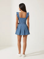 Model wearing a short-sleeved denim mini dress is crafted from a medium-blue wash denim featuring a structured, square-necked bodice with molded, bustier-style seaming and playful ruffled shoulder straps. Designed with a fitted waist cinched by a matching fabric belt with a metallic buckle, the dress flows down into a flared, pleated skater skirt that creates a crisp, youthful silhouette.
