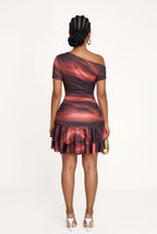 A woman viewed from the back wearing a one-shoulder ruched mini dress with a red, orange, and dark brown abstract swirl pattern. She is holding a small gold handbag and wearing white high heels, set against a plain white studio background.