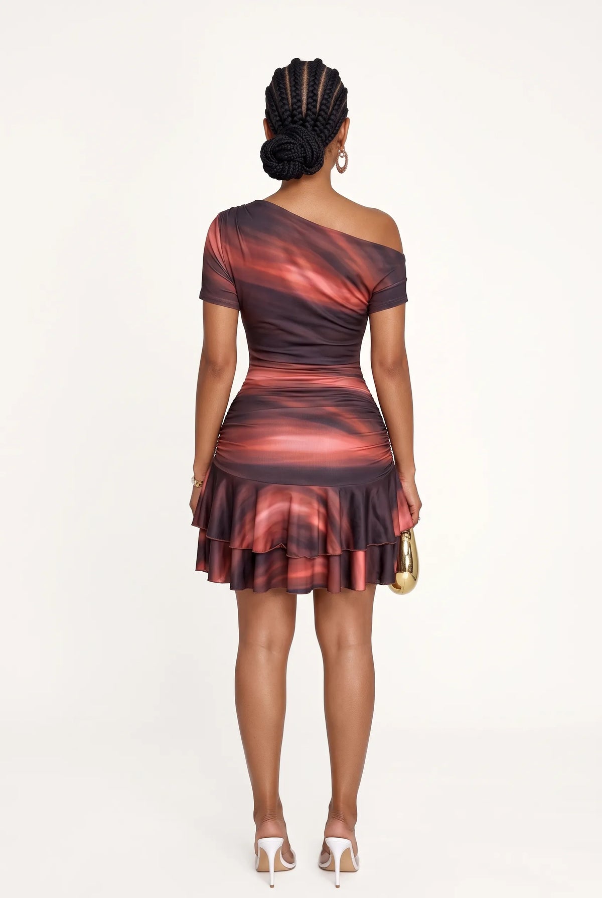 A woman viewed from the back wearing a one-shoulder ruched mini dress with a red, orange, and dark brown abstract swirl pattern. She is holding a small gold handbag and wearing white high heels, set against a plain white studio background.
