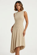 This sleeveless, beige midi dress features a crew neckline and a sophisticated sunray pin-tuck design that radiates outward across the fitted bodice. Crafted from a fluid, medium-weight fabric, the dress forms structural pleated lines that extend downward through an A-line silhouette, culminating in a dramatic asymmetrical high-low hemline that flows gracefully around the legs.