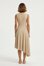 This sleeveless, beige midi dress features a crew neckline and a sophisticated sunray pin-tuck design that radiates outward across the fitted bodice. Crafted from a fluid, medium-weight fabric, the dress forms structural pleated lines that extend downward through an A-line silhouette, culminating in a dramatic asymmetrical high-low hemline that flows gracefully around the legs.