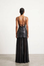 A model viewed from the back wearing a black strappy dress with a structured faux-leather bodice and a sheer black midi skirt, set against a minimalist off-white studio background.