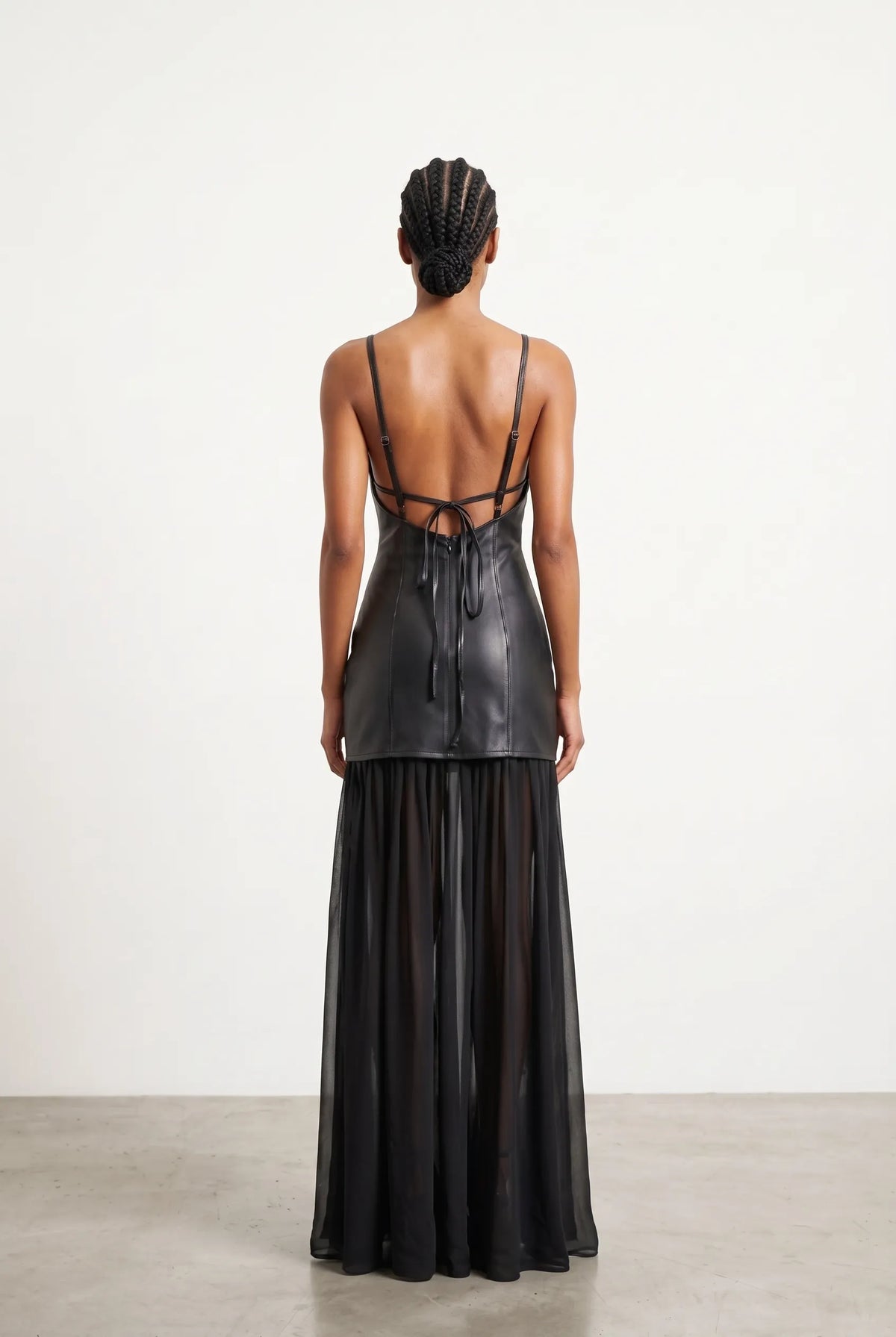 A model viewed from the back wearing a black strappy dress with a structured faux-leather bodice and a sheer black midi skirt, set against a minimalist off-white studio background.