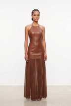 A model stands against a plain white studio background wearing a sleeveless brown faux-leather halter dress with a structured bodice and a flowing sheer pleated skirt.