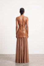 A model viewed from the back wearing a brown leather two-piece outfit consisting of a sleeveless wrap-style top with a tie-back and a matching pleated maxi skirt, set against a plain white studio background.
