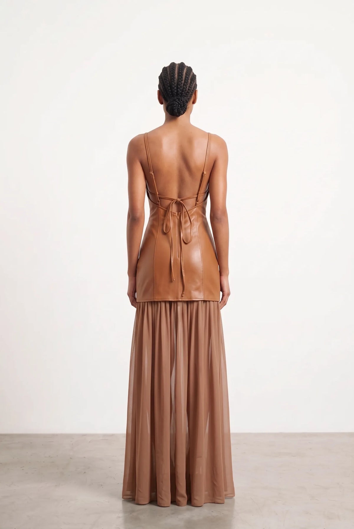 A model viewed from the back wearing a brown leather two-piece outfit consisting of a sleeveless wrap-style top with a tie-back and a matching pleated maxi skirt, set against a plain white studio background.