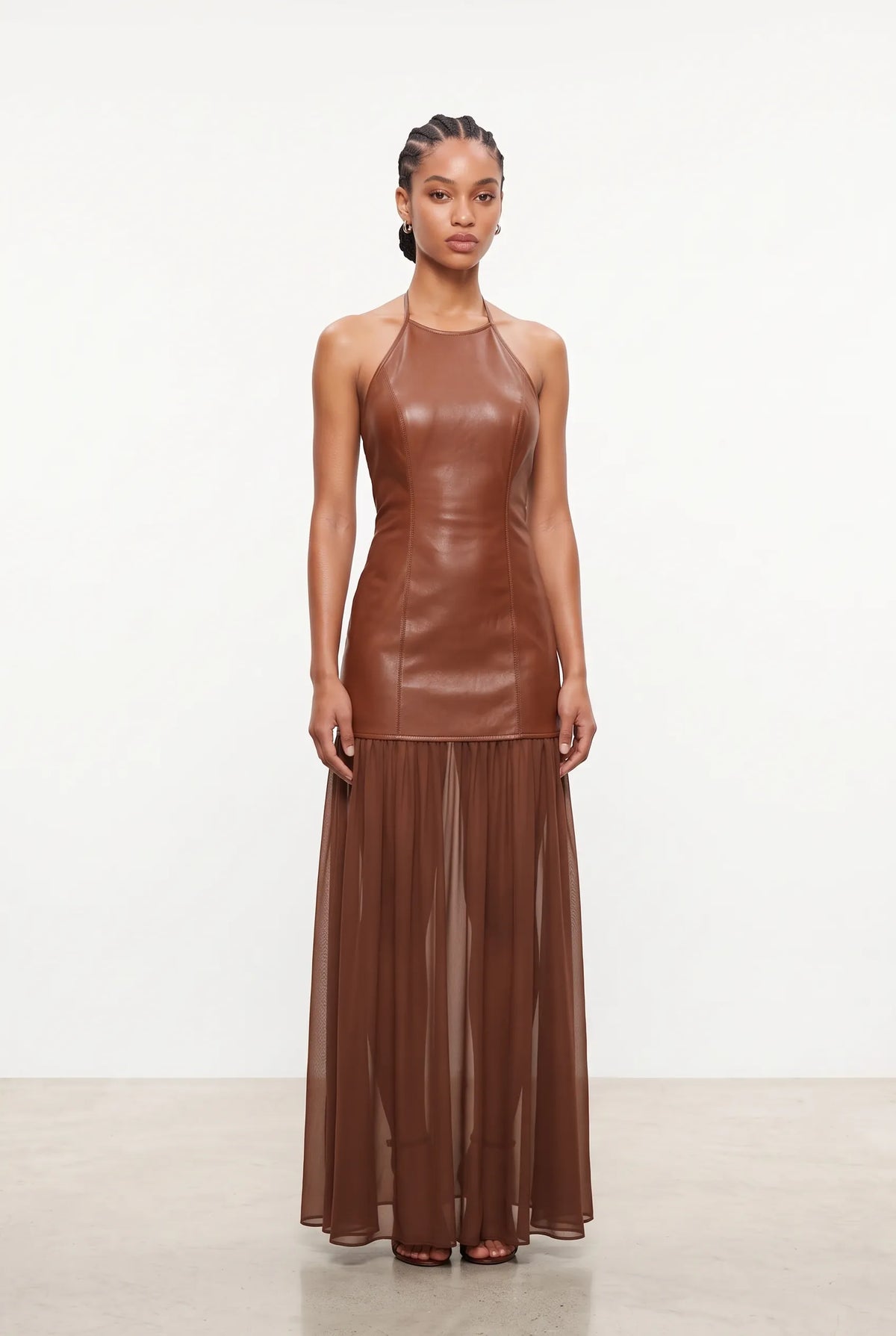 A model stands against a plain white studio background wearing a sleeveless brown faux-leather halter dress with a structured bodice and a flowing sheer pleated skirt.