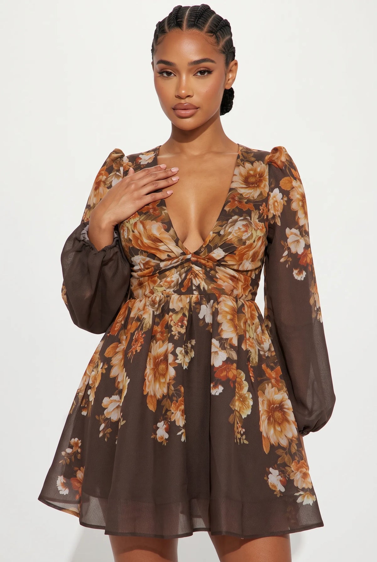 A woman modeling a brown long-sleeve mini dress with a deep V-neckline, twisted waist detail, and an orange, beige, and white floral print, set against a plain light gray background.