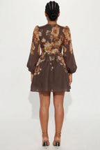 A woman viewed from the back wearing a brown sheer long-sleeve mini dress with a large floral print in beige, orange, and white, paired with brown strappy high heels against a plain white studio background.