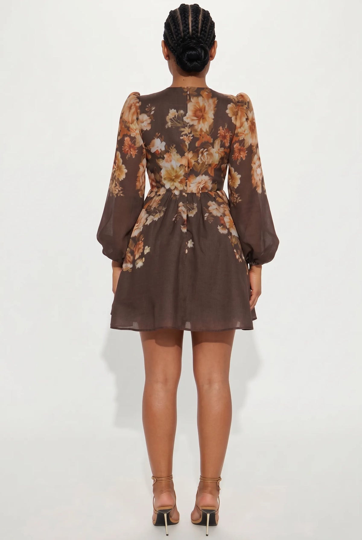 A woman viewed from the back wearing a brown sheer long-sleeve mini dress with a large floral print in beige, orange, and white, paired with brown strappy high heels against a plain white studio background.