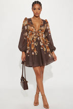 A woman modeling a brown long-sleeve mini dress with a floral print in orange, beige, and white, featuring a deep V-neck, puff sleeves, and a tie at the waist. She is holding a small brown handbag and wearing brown strappy high-heeled sandals against a plain white studio background.
