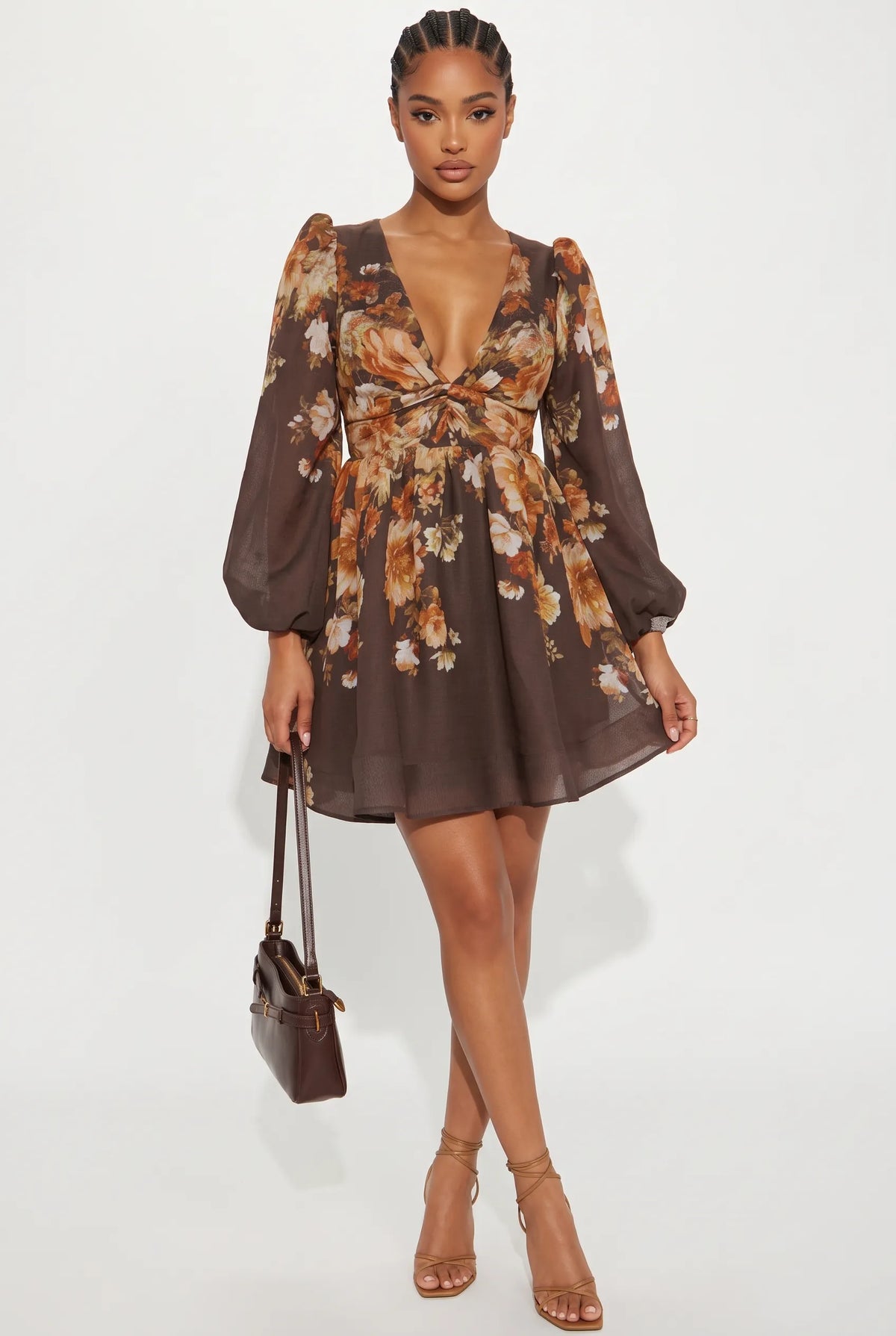 A woman modeling a brown long-sleeve mini dress with a floral print in orange, beige, and white, featuring a deep V-neck, puff sleeves, and a tie at the waist. She is holding a small brown handbag and wearing brown strappy high-heeled sandals against a plain white studio background.