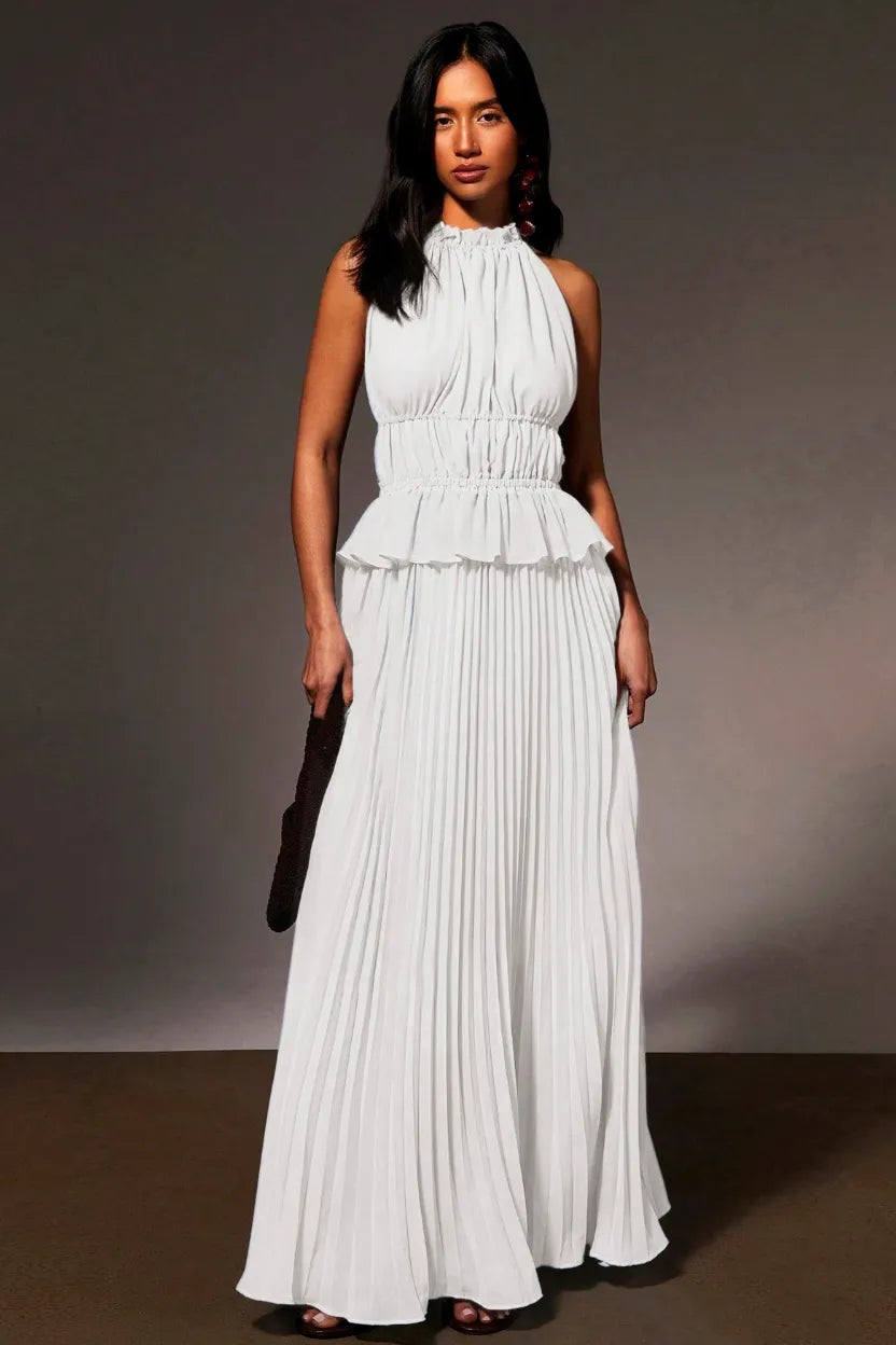 A woman modeling a sleeveless white halter-neck maxi dress with a gathered peplum waist and pleated skirt, standing against a dark gray studio backdrop.