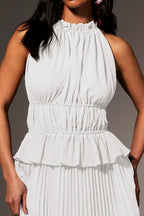 A close-up of a model wearing a white sleeveless halter-neck dress with a ruffled neckline, a gathered smocked waist, and a pleated skirt, set against a solid gray background.