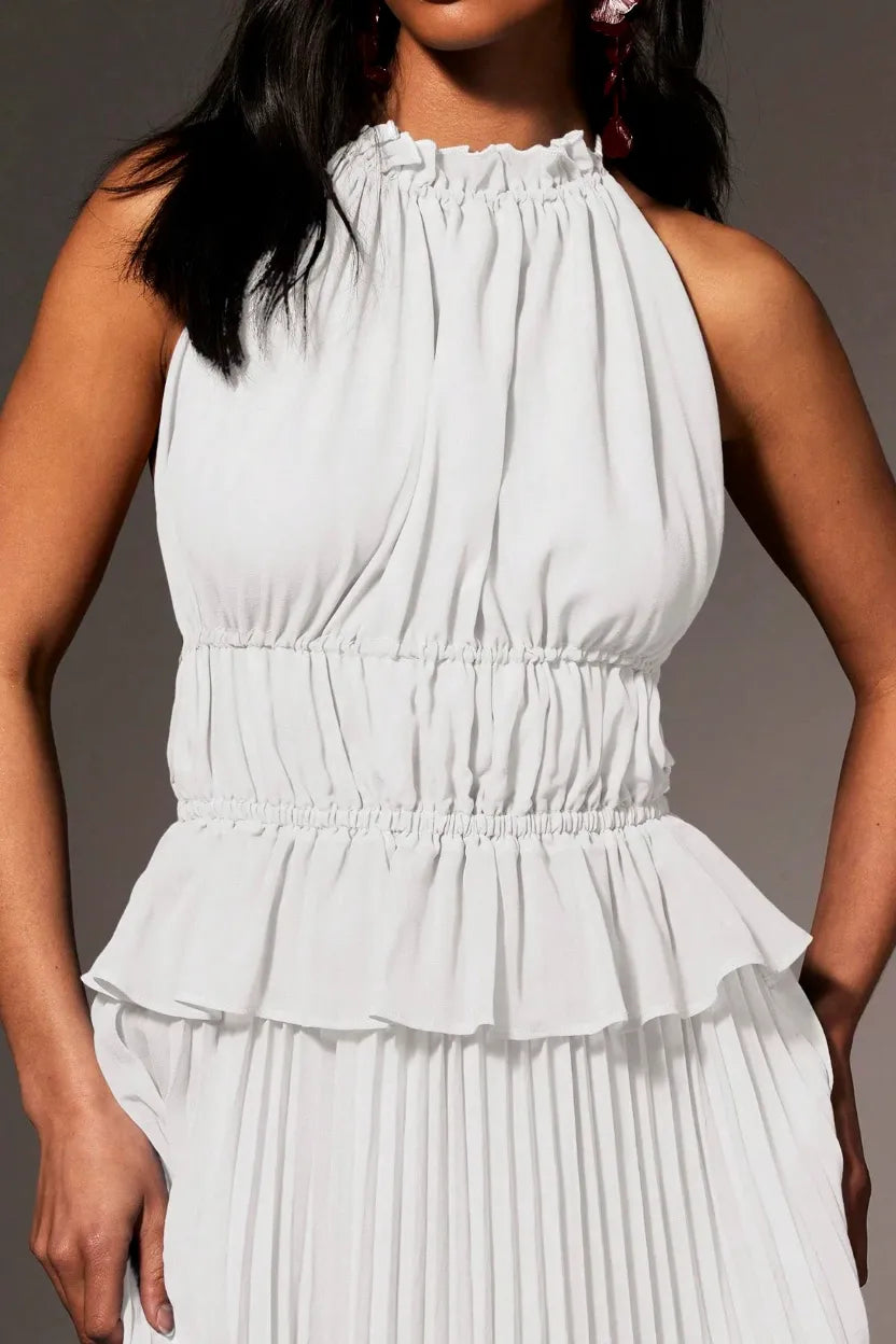 A close-up of a model wearing a white sleeveless halter-neck dress with a ruffled neckline, a gathered smocked waist, and a pleated skirt, set against a solid gray background.