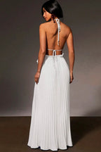 A woman shown from the back wearing a white sleeveless maxi dress with a pleated skirt, tie-back straps, and an open back, photographed against a dark gray studio background.