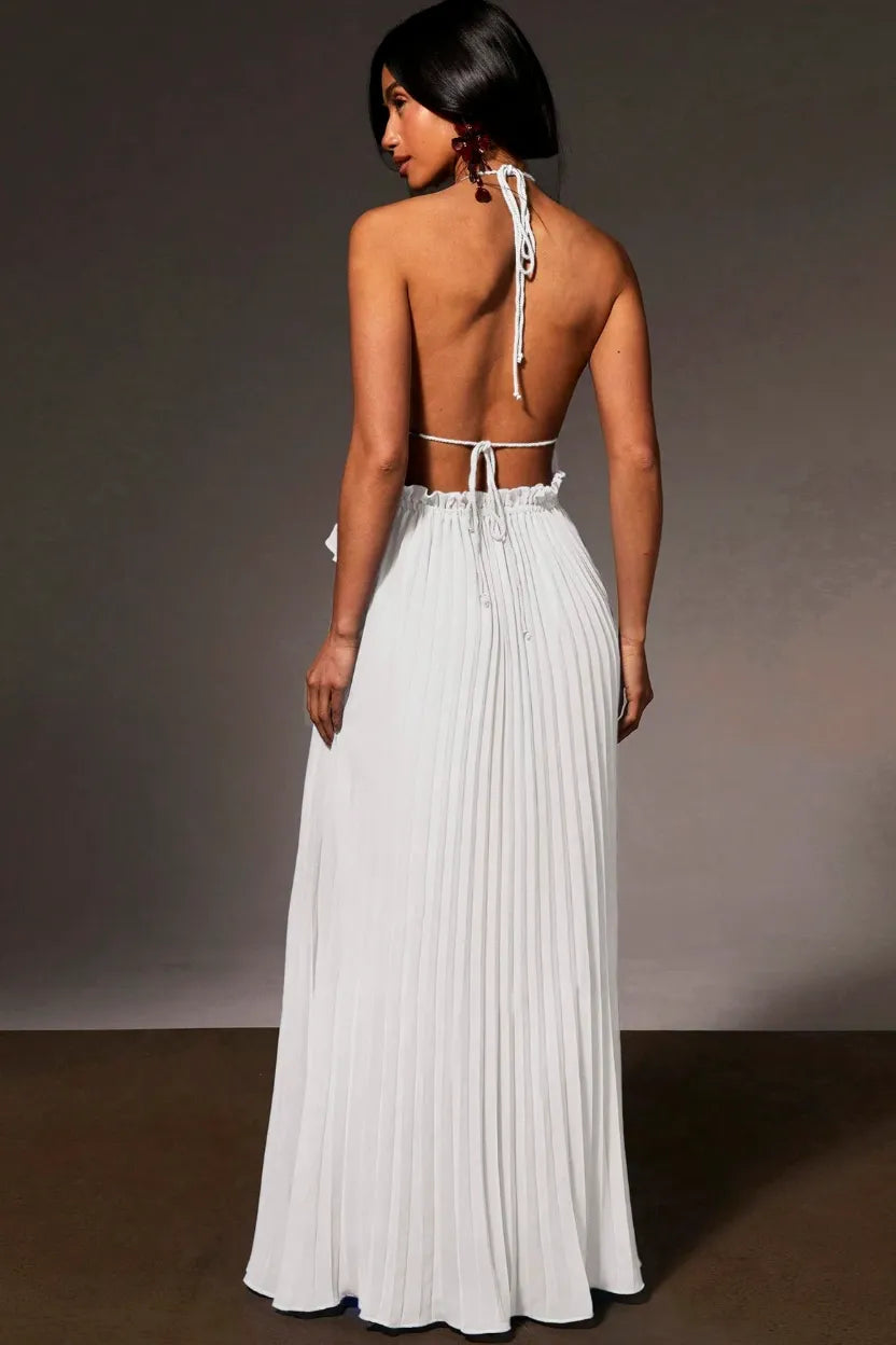A woman shown from the back wearing a white sleeveless maxi dress with a pleated skirt, tie-back straps, and an open back, photographed against a dark gray studio background.