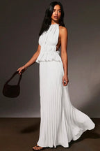 A woman modeling a white halter-neck maxi dress with a ruffled peplum waist and side cutouts, holding a dark brown handbag against a dark gray studio background.