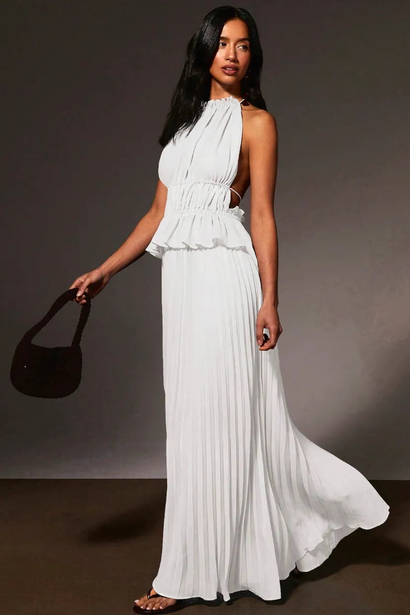 A woman modeling a white halter-neck maxi dress with a ruffled peplum waist and side cutouts, holding a dark brown handbag against a dark gray studio background.