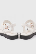 A pair of white T-strap flat sandals with black platform soles and rose gold buckle details, shown from a rear three-quarter angle on a light gray background.