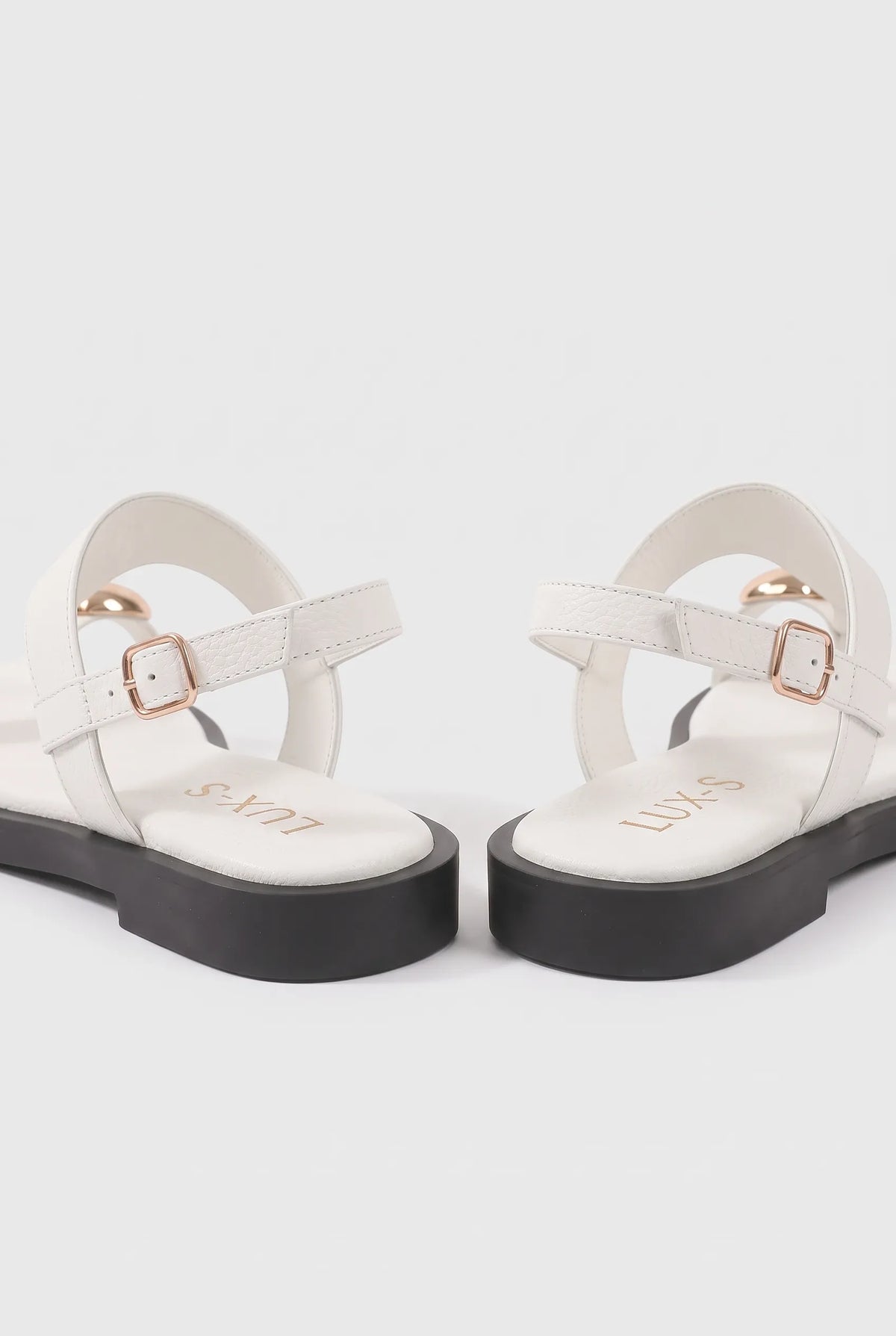 A pair of white T-strap flat sandals with black platform soles and rose gold buckle details, shown from a rear three-quarter angle on a light gray background.