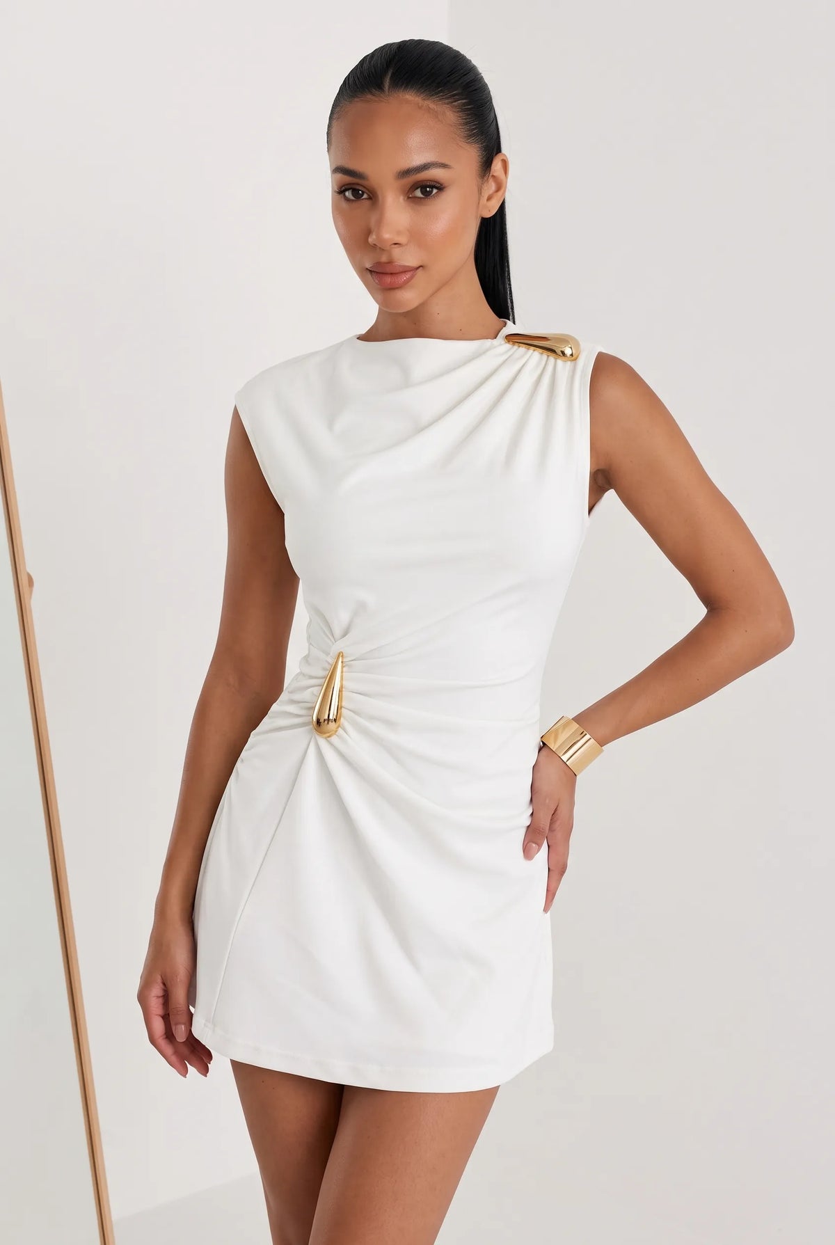 A woman modeling a white one-shoulder ruched mini dress with gold metallic accents on the shoulder and waist, set against a plain light gray studio background.