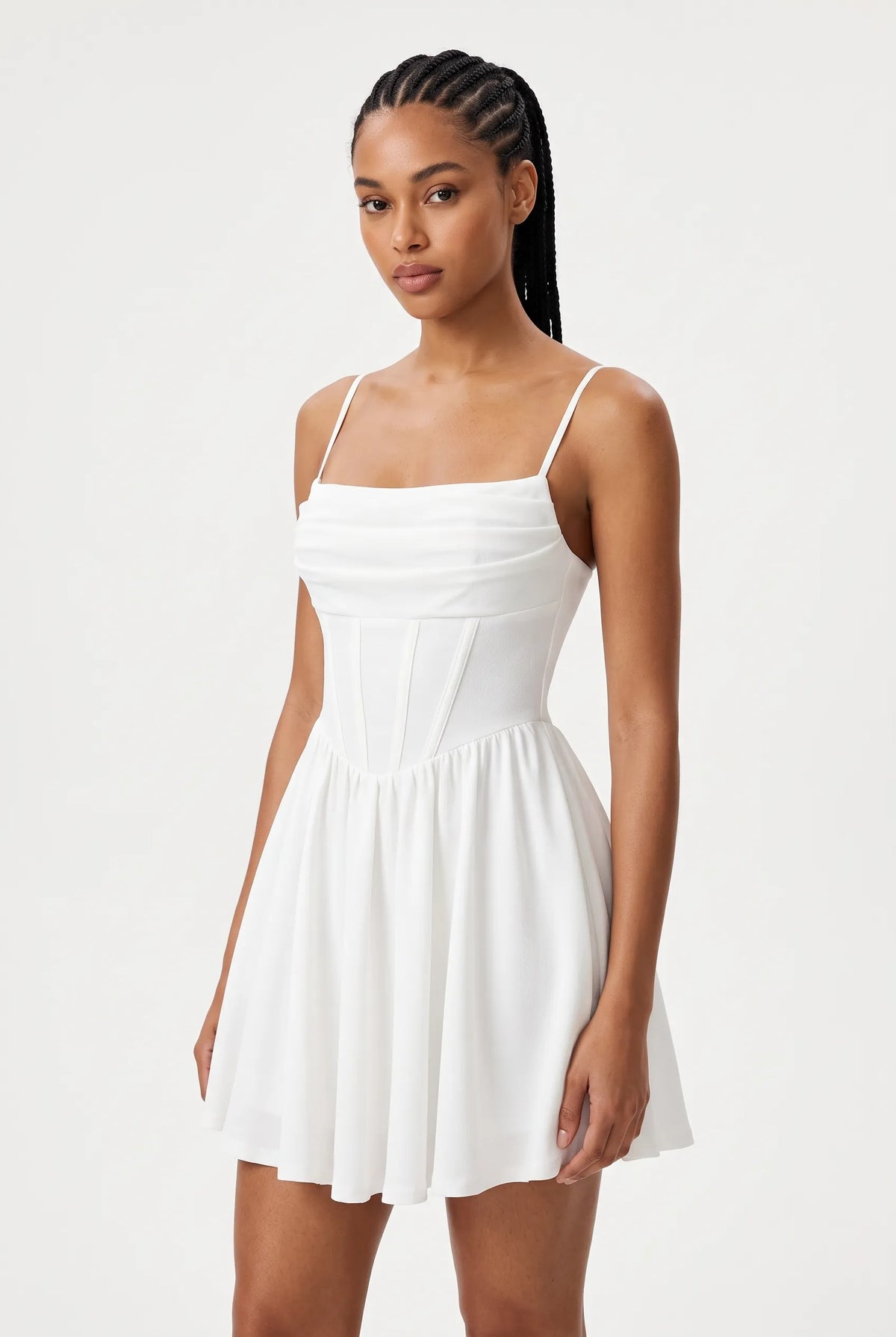 A woman modeling a white spaghetti-strap mini dress with a fitted bodice and flared skirt, photographed against a clean light gray studio background.